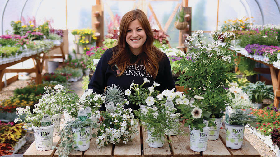 Laura Garden Answer stands by a table brimming with diverse plants highlighting her passion for gardening